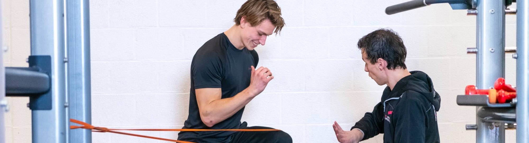Student en docent oefenen in gymzaal, jongen zit op 1 knie en stretcht zijn been.