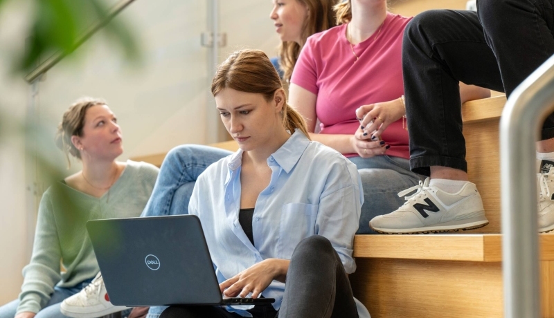 Studenten zittend op de trap in de hal van gebouw R26, werken op de laptop.