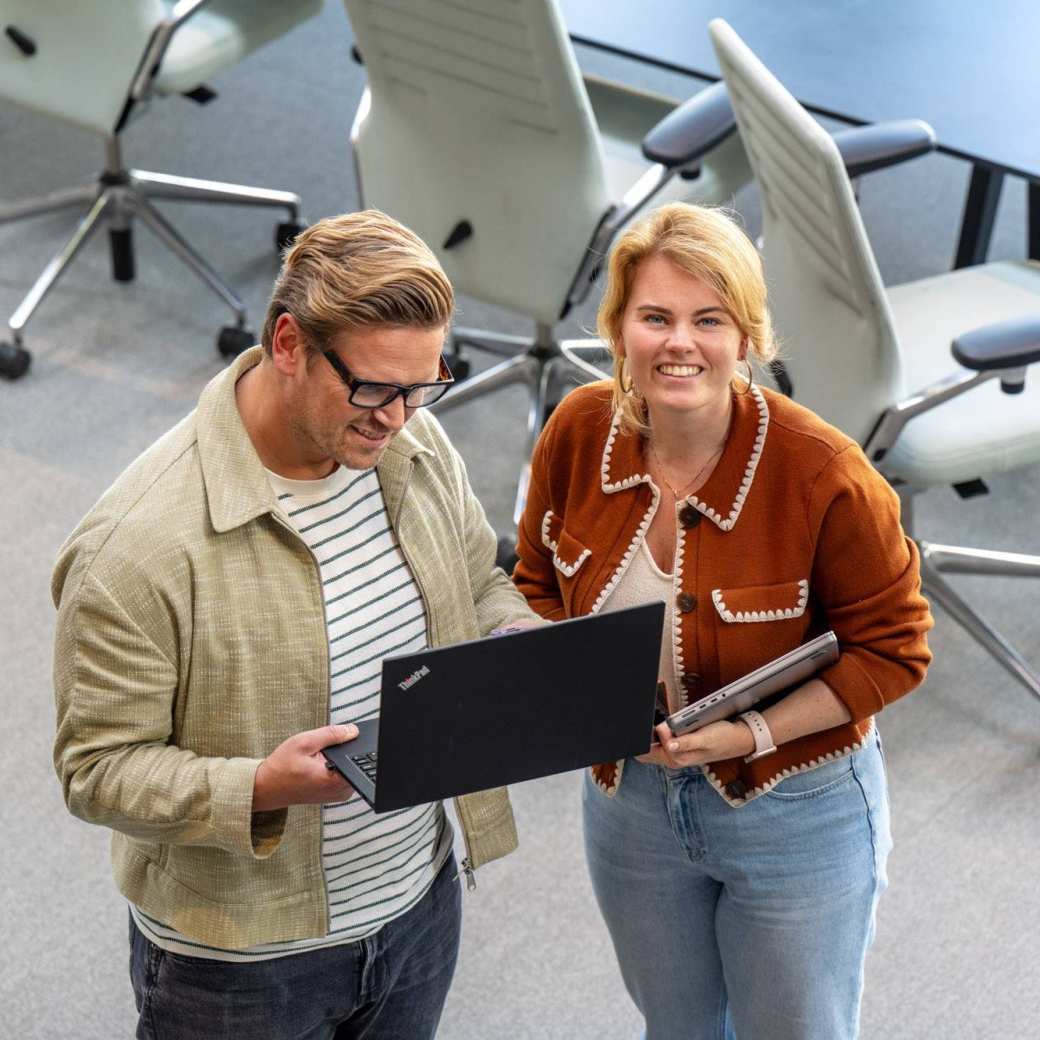 Man en vrouw staan met laptop in de hand en kijken lachend naar camera