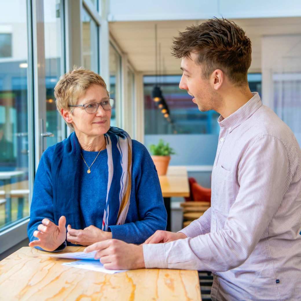 Man en vrouw staan aan een tafel bij een raam te praten.