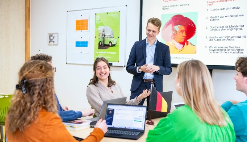 Studenten zitten samen aan tafels met laptops en boeken in het klaslokaal. De leraar staat ervoor en praat met hen mee.
