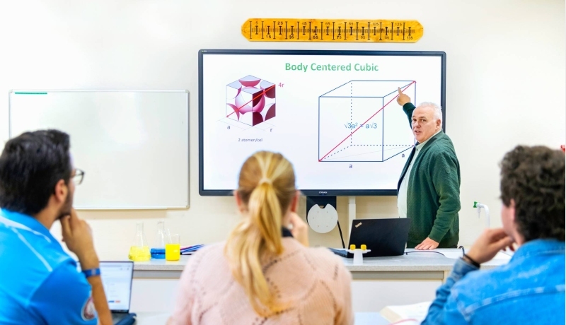Drie natuurkundecursisten luisteren naar een docent bij het whiteboard.
