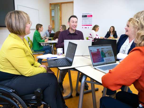 Een groepje van vier mensen in gesprek met laptops op tafel in een lokaal met anderen