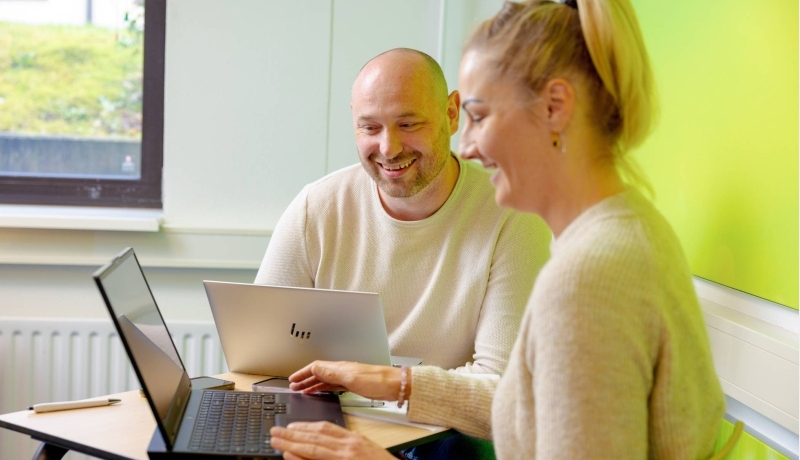 Vrouw en man zitten achter laptop, allebei aan het lachen. Groene muur op achtergrond.