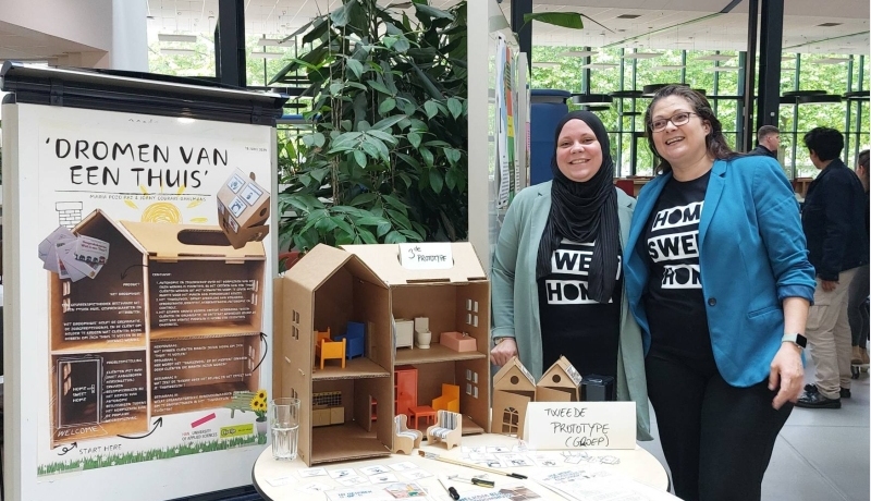 Studenten Joany en Marie staan voor een tafel waarop een soort poppenhuis te zien is. Op de poster naast hen staat informatie over een droomhuis.