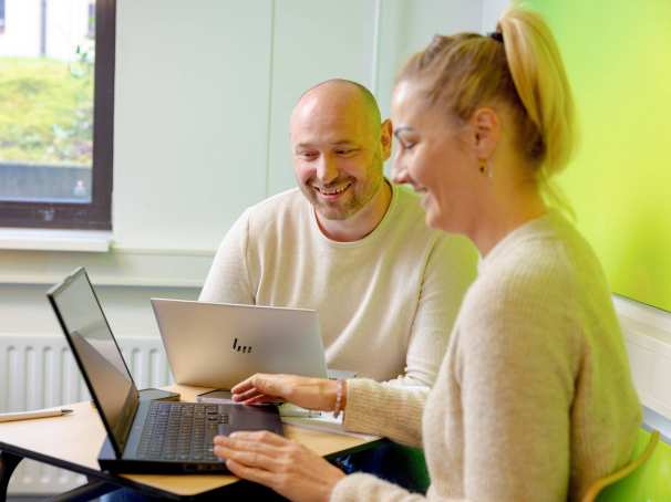 Vrouw en man zitten achter laptop, allebei aan het lachen. Groene muur op achtergrond.