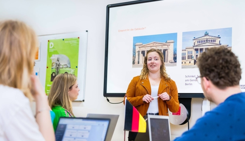 Docent geeft aan twee studenten uitleg en wijst daarbij naar de pagina in het boek dat de studenten in de hand hebben.