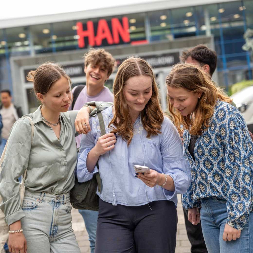 Studenten staan met een groepje buiten bij de ingang van gebouw R26, kijken op een telefoon.