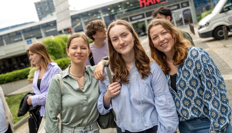 Studenten staan met vriendengroep buiten bij de ingang van gebouw R26, kijken in de camera.