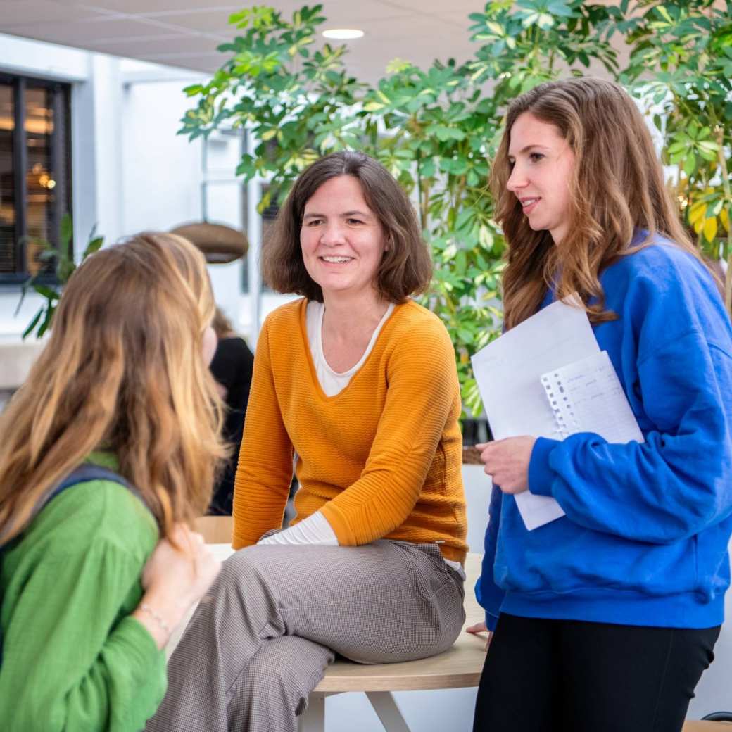 3 studenten gekleed in oranje, blauw en groen, overleggen met elkaar op de gang.