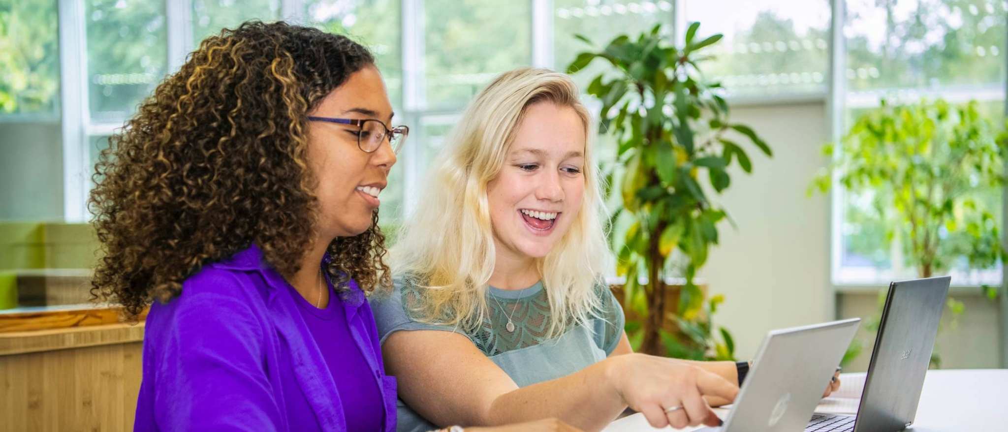 Twee studenten Leraar Wiskunde zitten naast elkaar met een laptop. De ene wijst lachend op het scherm iets aan en de ander kijkt lachend mee.
