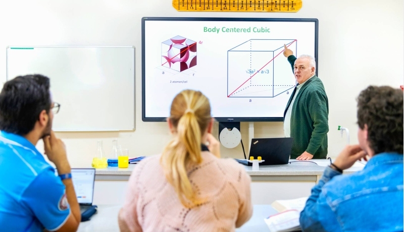 Drie natuurkundecursisten luisteren naar een docent bij het whiteboard.