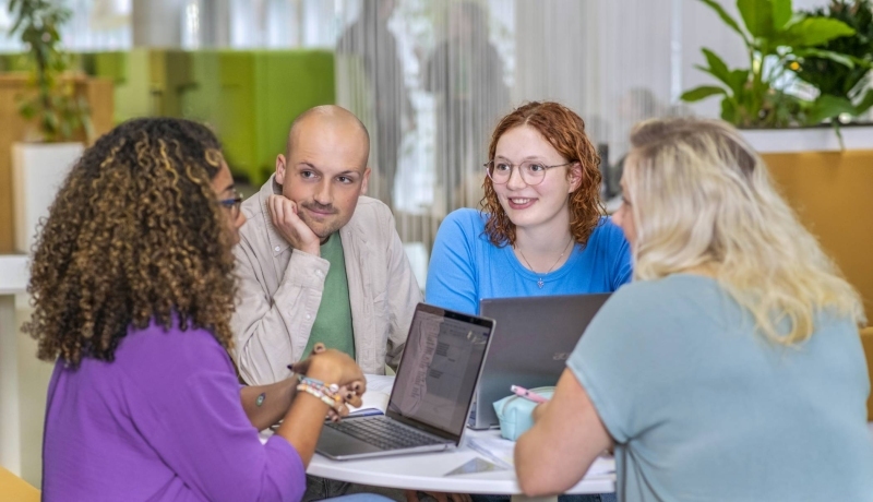 Vier studenten Leraar Wiskunde overleggen aan een ronde tafel en luisteren aandachtig naar elkaar. Op de tafel liggen etuis, schriften en laptops.