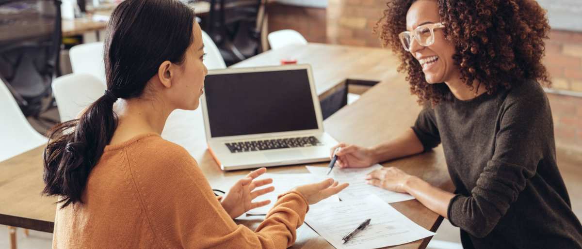 Twee vrouwen in gesprek in een kantoorsetting. Ze lachen en hebben een laptop en papieren.