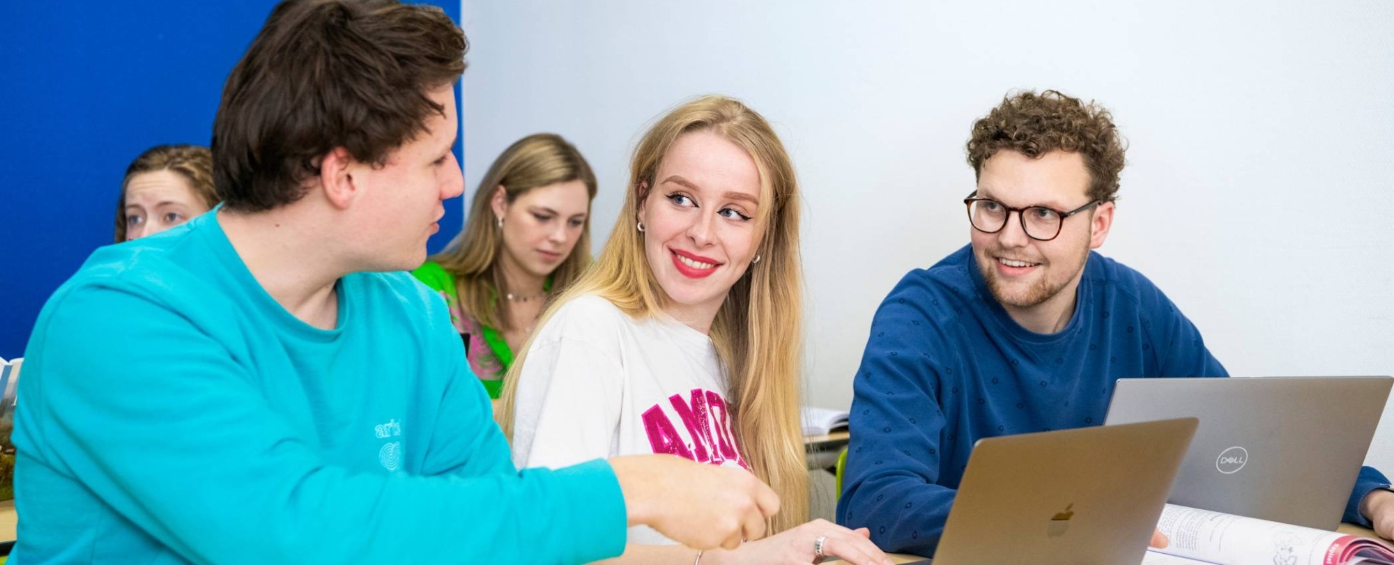 Drie studenten zitten op een rijtje in het klaslokaal met hun laptops opengeklapt. Ze zijn aan het overleggen.