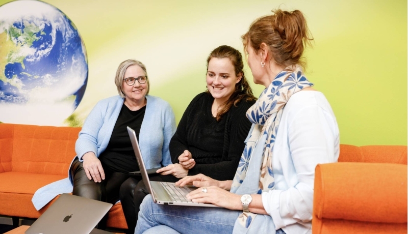 vrouw wijst iets aan op laptop, oranje banken. Wereldbol op achtergrond, tweede laptop op tafel.