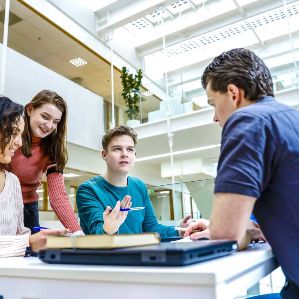 Drie studenten zitten te werken en één student stelt een vraag aan de docent die aandachtig luistert.