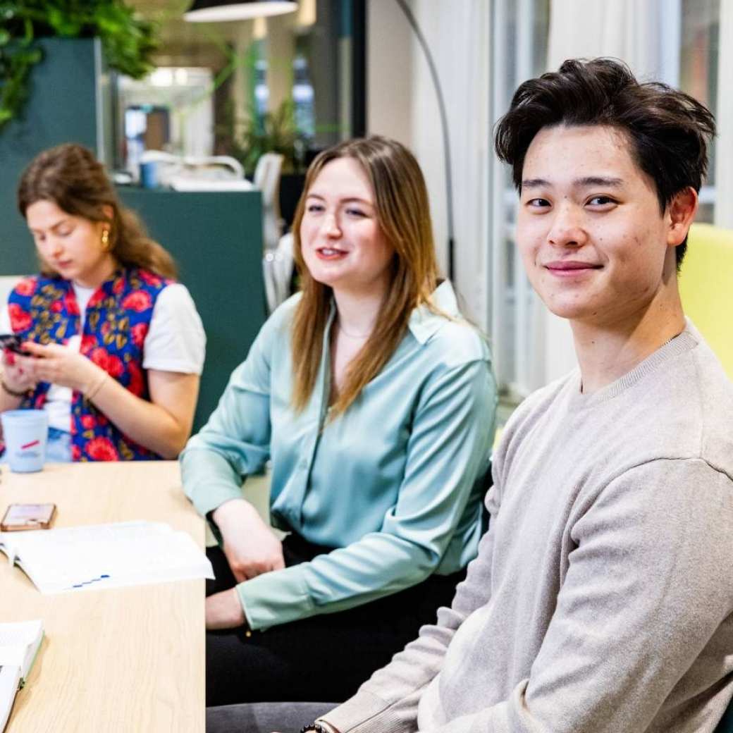 Bedrijfskunde studenten zitten aan een tafel en één student kijkt in de camera