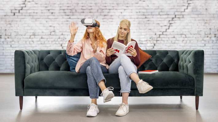 Een meid met vr-bril en een meid met een boek zitten samen op de bank