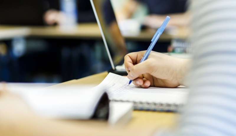 een hand met pen schrijft op een kladblok. erachter staat een laptop en op de voorgrond liggen wat papieren