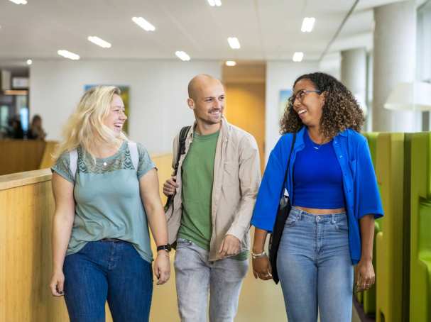 Drie studenten Leraar Wiskunde lopen lachend naast elkaar op de gang in de school