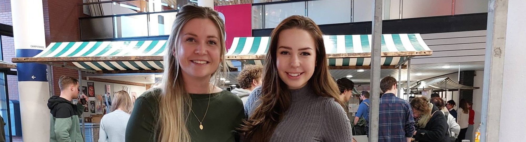 Hanne Wanink heeft blond haar en een groene top en draagt een gouden ketting. Romy Burghouts heeft een grijze jurk aan en donkere haren. Ze houden een poster in hun hand van het project met daarop Thuiszitten, dat is pas duur.