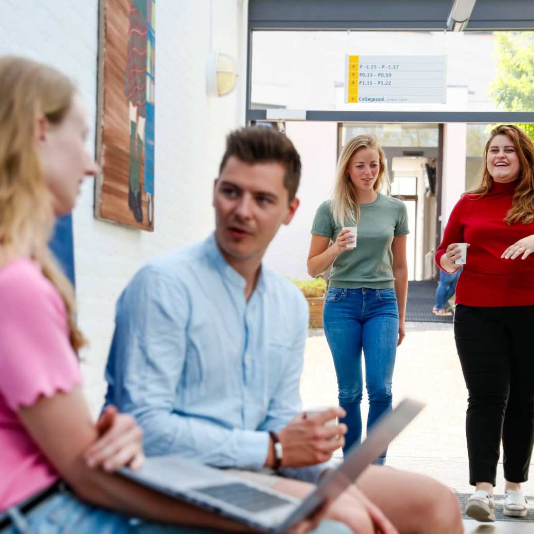 Een masterstudent zit op de rand van de tafel met een laptop in z'n hand en legt wat uit aan de rest.
