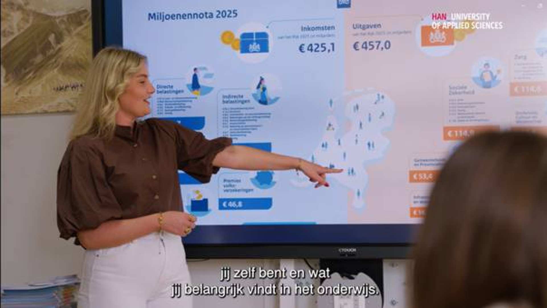 Student Leraar Economie wijst iets aan op het bord. Heeft kort haar, blauw/bruine blouse en een witte broek
