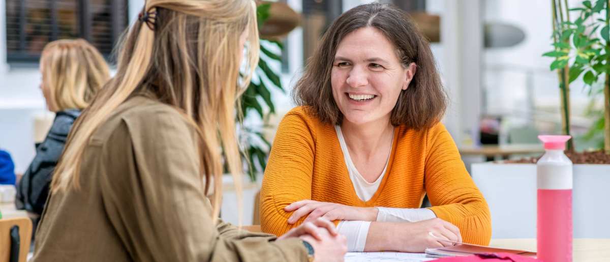 Vrouw in oranje trui lacht naar medestudent.