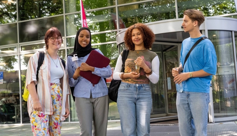 Vier studenten staan bij gebouw LvS10 bij draaideur, buiten. Op een rijtje. 