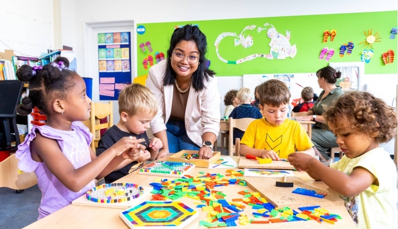 Kinderen leggen een puzzel in de klas en de lerares van de opleiding leraar basisonderwijs helpt hen hierbij
