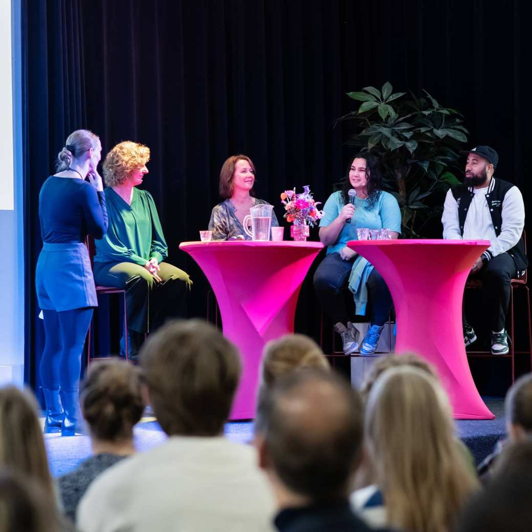 Podium met daarop een tafel met een rood-roze kleed. Er zitten meerdere deelnemers aan een panel achter de tafel. Een vrouw met blonde krullen en groene blouse, Natascha, een vrouw met halflang rood haar, Lisbeth, een jonge vrouw met blauwgroen shirt met lange mouwen en zwarte krullen en een jongen met een pet, Melvin.