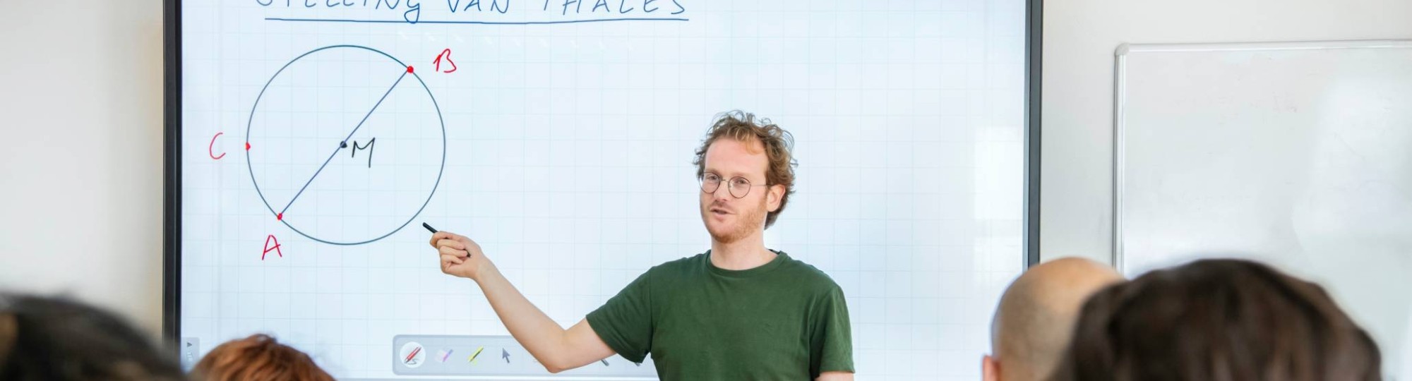 Leraar Wiskunde kijkt naar zijn klas, terwijl zijn hand wijst naar de cirkel op het bord waar 'stelling van Thales' boven staat