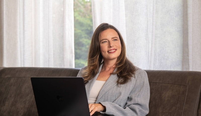 Vrouw in grijs pak zit op de bank met haar laptop en lacht