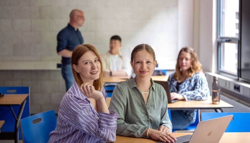 Twee studenten zijn samen aan het werk op de laptop, kijken in de camera, zijn in een klas. 