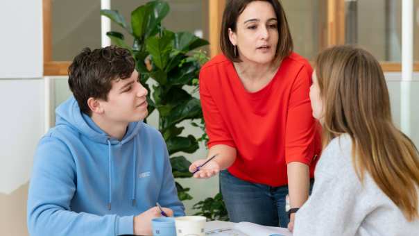 Docent met rode trui geeft uitleg aan twee studenten