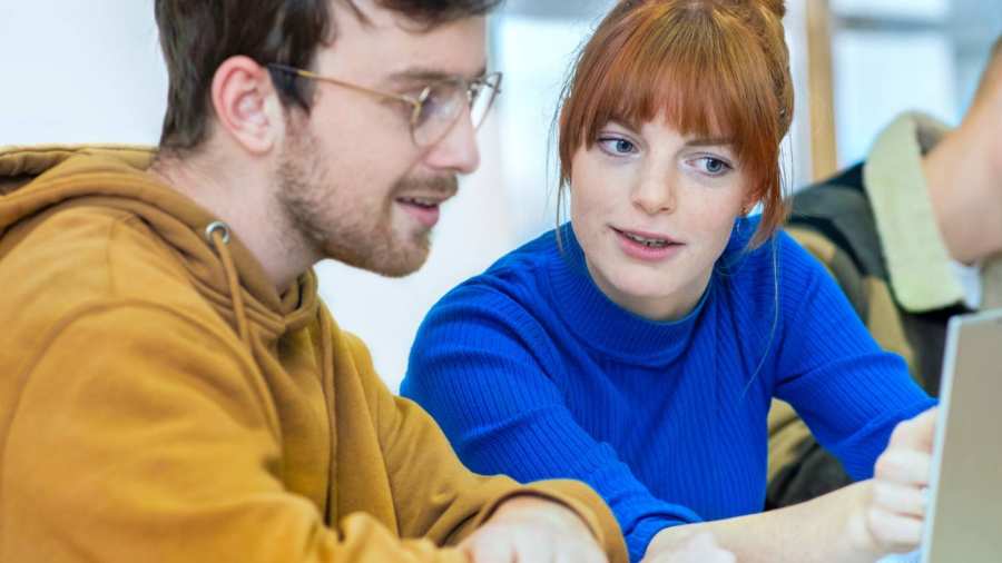 Twee studenten zitten naast elkaar en kijken samen aandachtig naar de laptop die voor hun staat.
