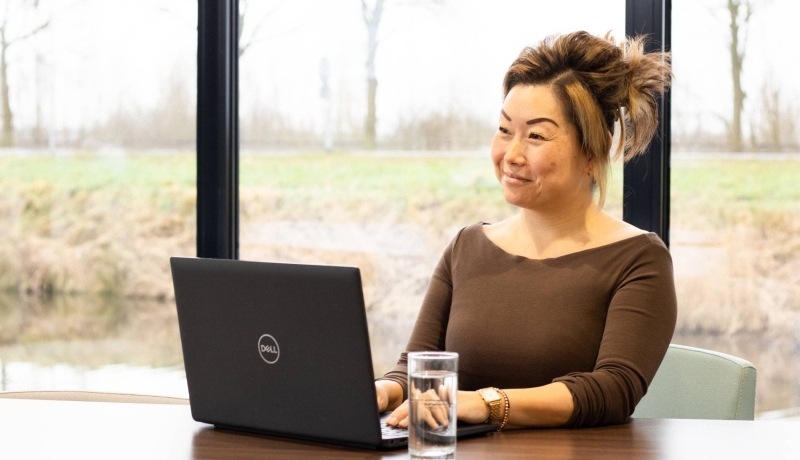 Dame poseert met een glimlach, achter een laptop. Een glas water staat naast haar op het bureau.