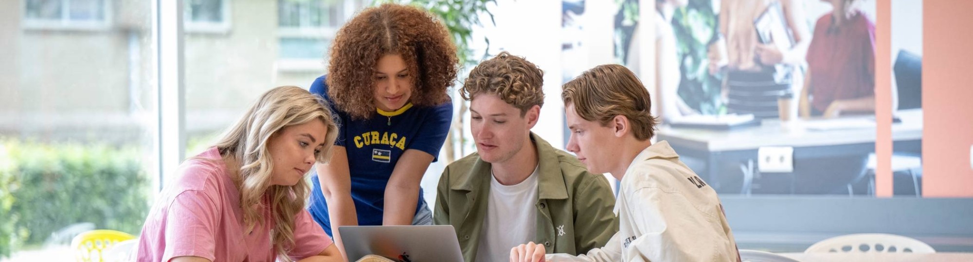 Groepje studenten werken samen aan een ronde tafel in de gang, werkplek in LvS10. Kijken samen naar de laptop.