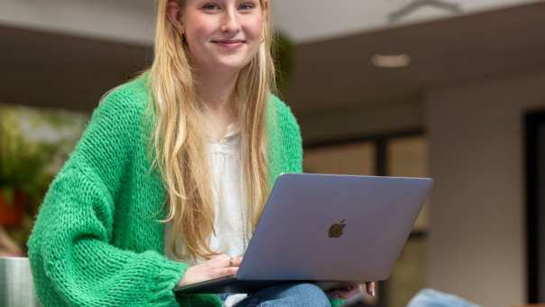een student/meisje met een groen vest en lang blond haar zit met een laptop op schoot en kijkt lachend in de camera.