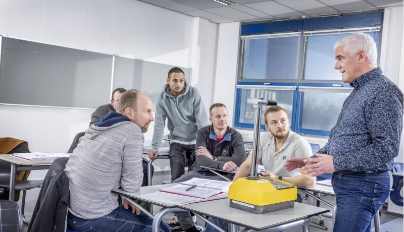 5 mannen zittend en staand aan een tafel luisteren aandachtig naar de docent