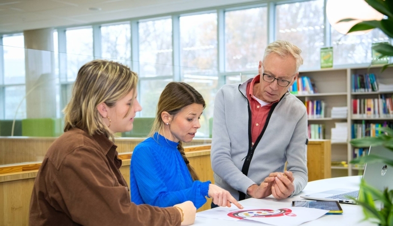 Een man en twee vrouwen in gesprek aan een tafel. Een vrouw wijst iets aan op papier.