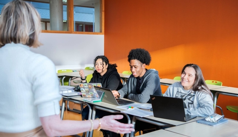 Docent zit op een tafel en geeft uitleg aan 3 masterstudenten die lachen