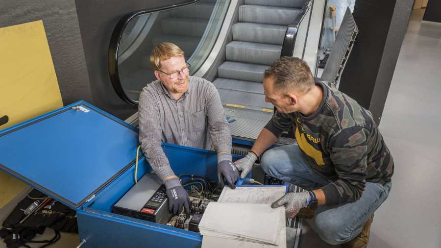 2 mannen, vastgoedinspecteurs, zitten onderaan een roltrap en zijn in gesprek over wat er met de roltrap aan de hand is.