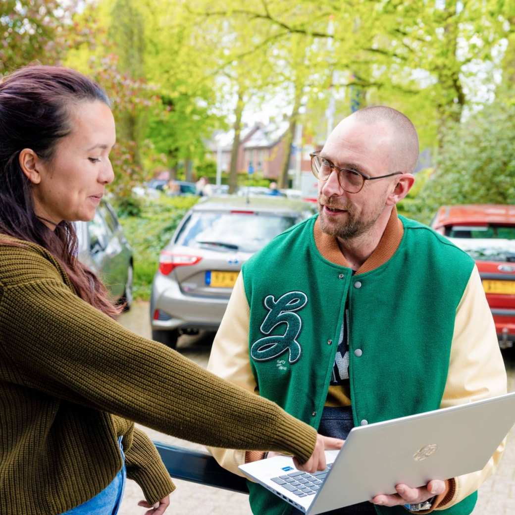 Vrouw wijst iets aan op laptop buiten, man heeft groen jack aan. Vrouw heeft groene trui aan.