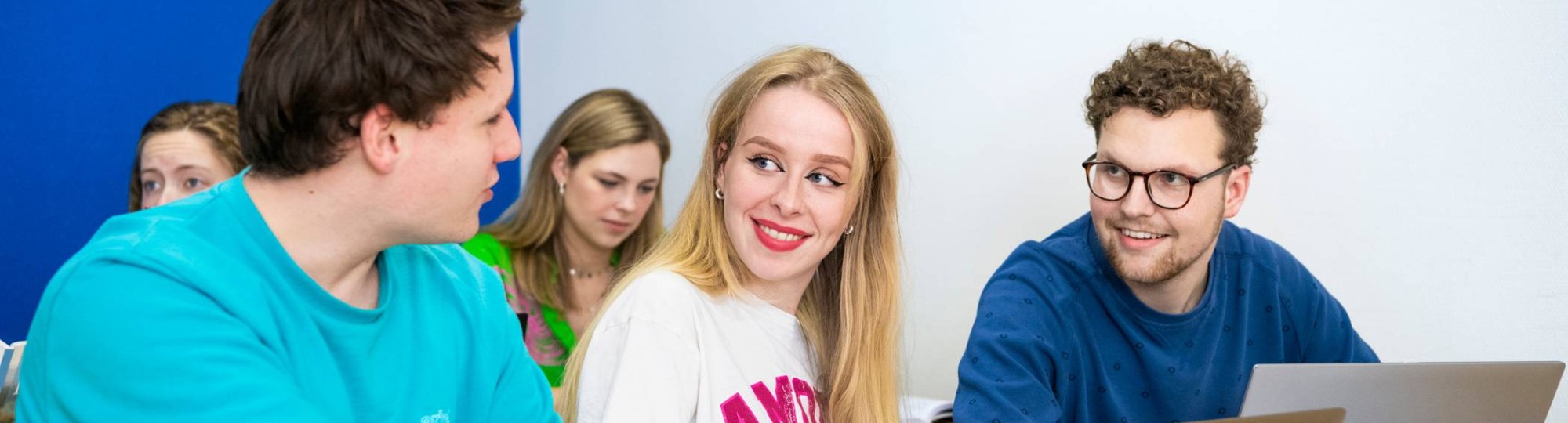 Drie studenten zitten op een rijtje in het klaslokaal met hun laptops opengeklapt. Ze zijn aan het overleggen.