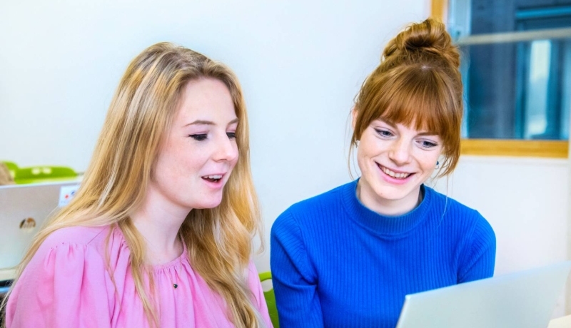 Twee vrouwelijke studenten leraar Geschiedenis zitten naast elkaar en kijken lachend naar het scherm (laptop) dat voor hun staat.