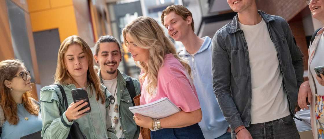 Groepje studenten staan bij elkaar en hebben plezier. Studenten kijkt op haar telefoon, ze staan in de gang. 
