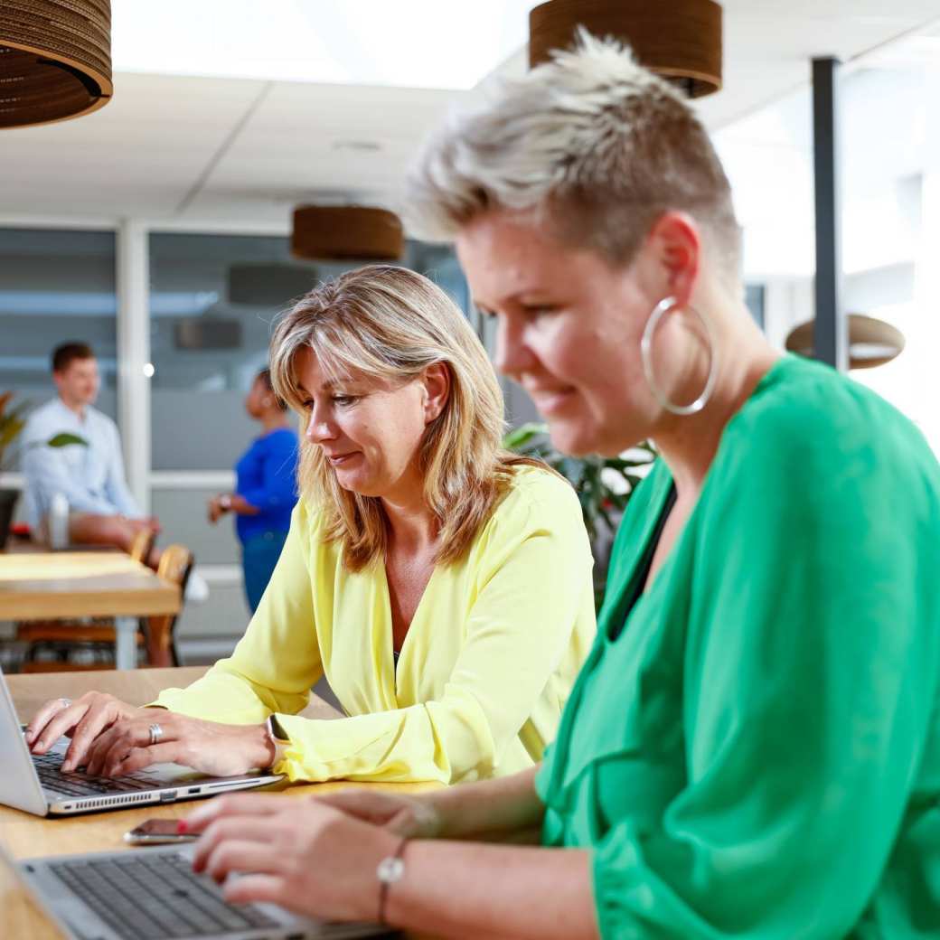 Een vrouw met een gele trui en een vrouw met een groene trui achter de laptop