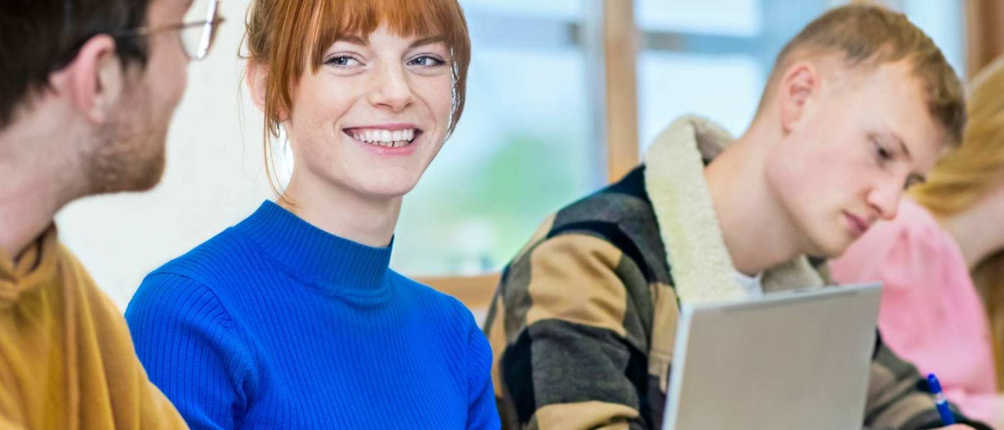 Vier studenten zitten op een rij. Een student specifiek is scherp in beeld en ze lacht naar de persoon naast haar.